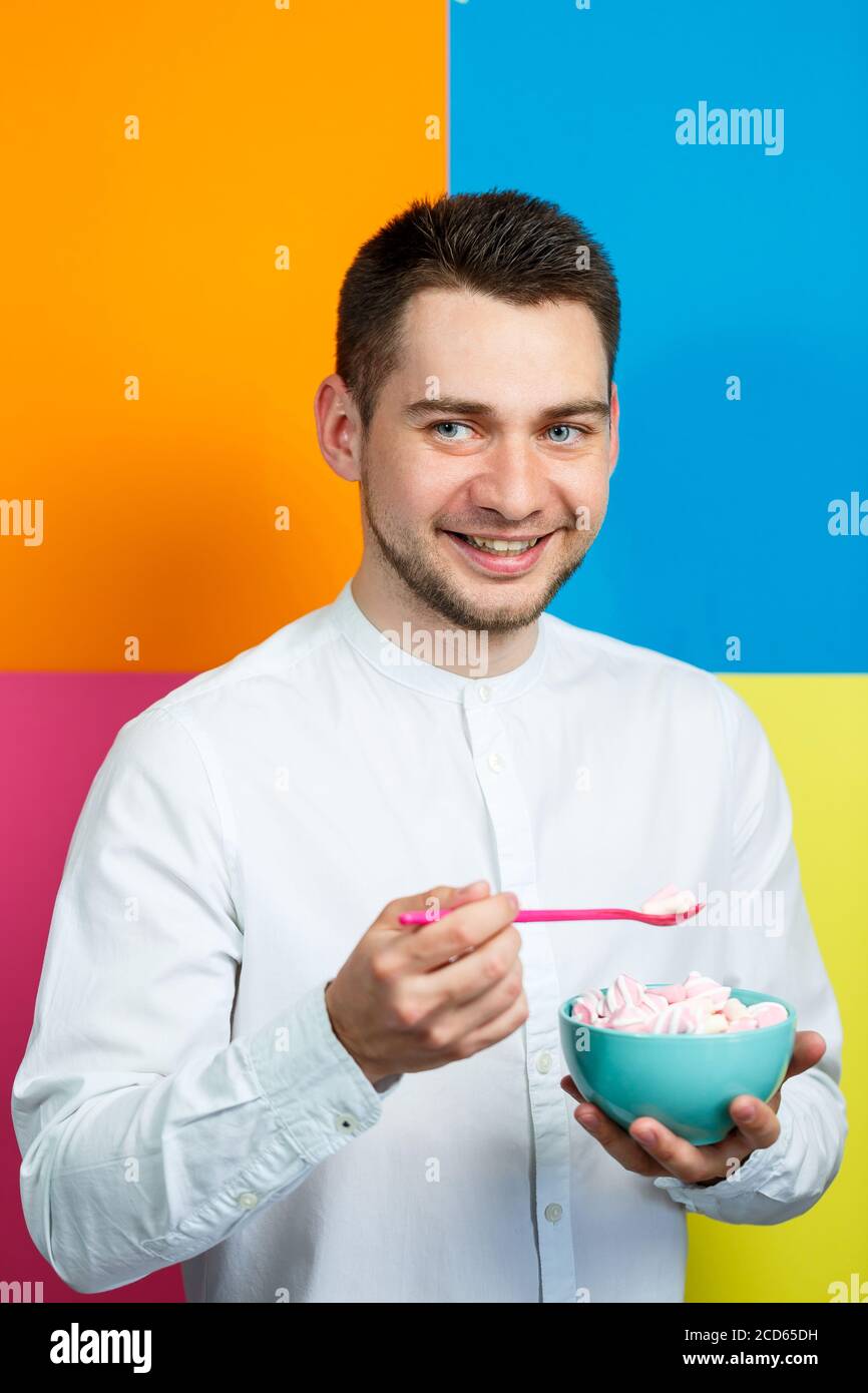 Un homme sur fond jaune, bleu, orange, rose dans une chemise blanche avec un plateau turquoise de guimauves et une cuillère rose. Le gars avec un sourire. Un im Banque D'Images