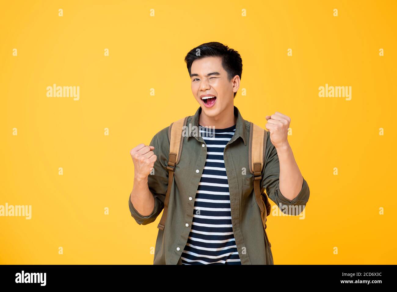 Portrait d'un heureux enthousiaste jeune homme de routard asiatique enthousiaste élevant ses poings faisant le geste de succès dans le studio isolé fond jaune Banque D'Images