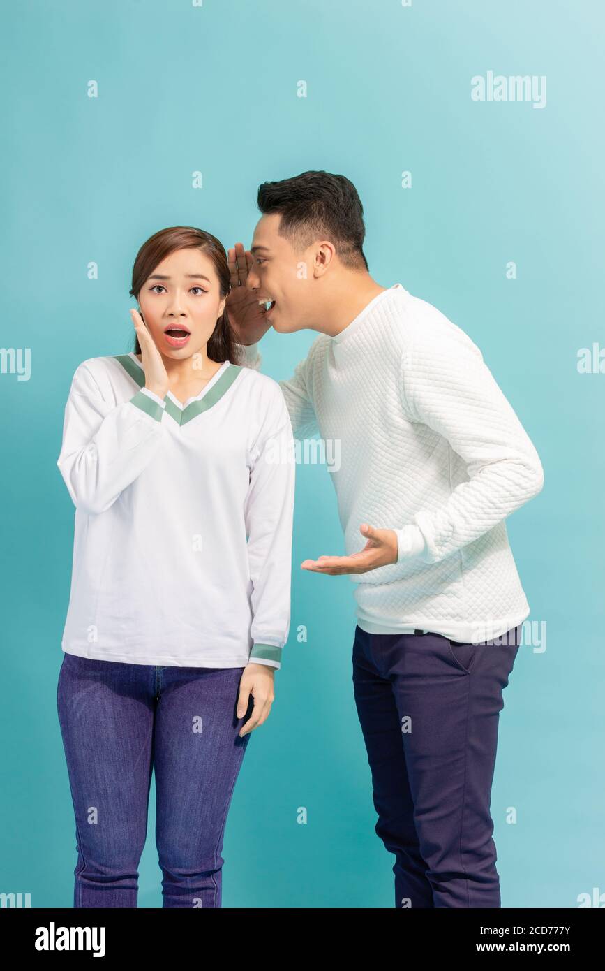 Homme asiatique partageant un secret ou chuchotant des gossips dans l'oreille de sa petite amie, fond de studio bleu Banque D'Images