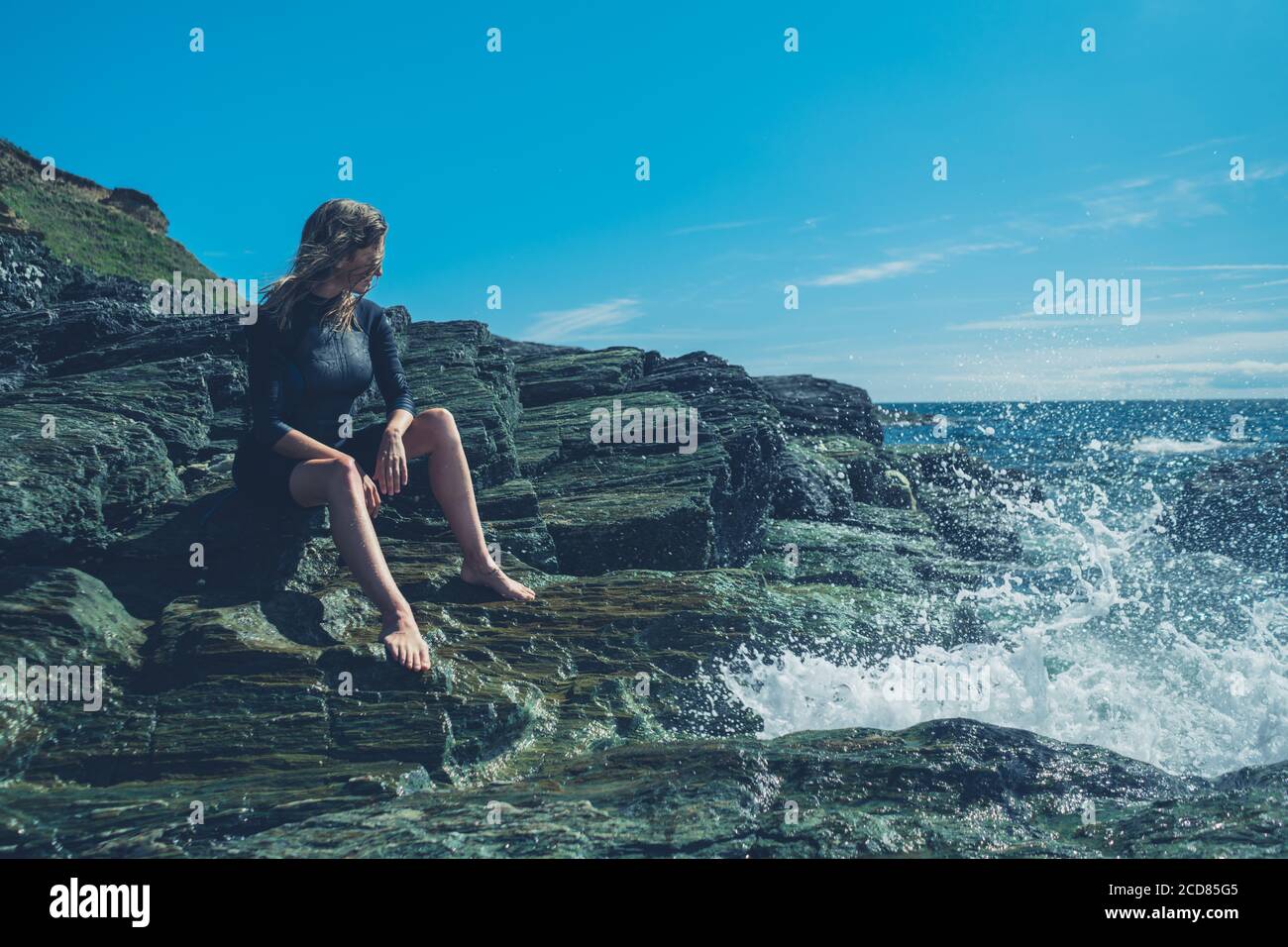 Une jeune femme portant une combinaison est assise sur le rochers au bord de la mer tandis que les vagues s'écrasant Banque D'Images