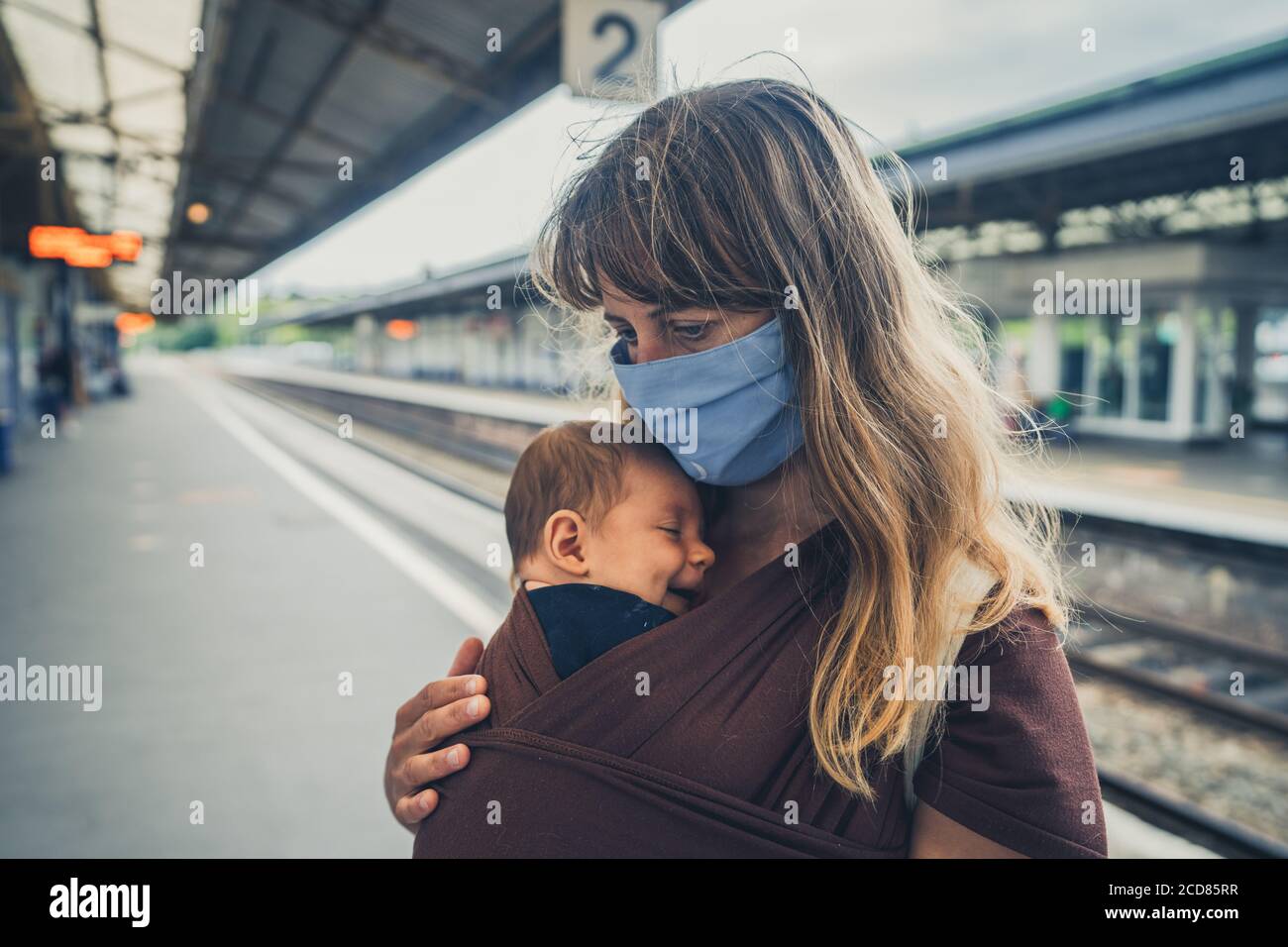 Une jeune mère avec son bébé dans une élingue est port d'un masque facial à une gare Banque D'Images