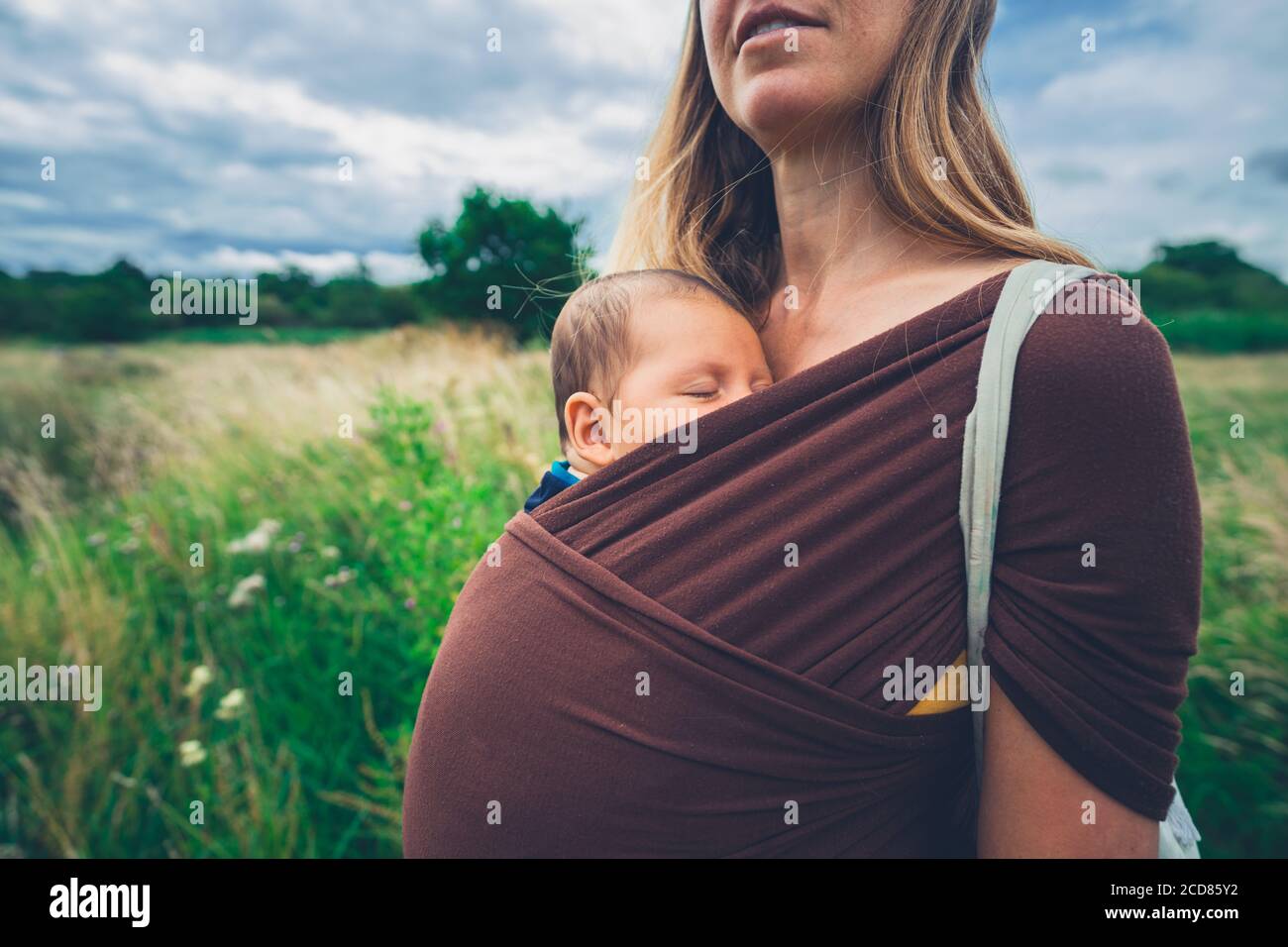 Une jeune mère avec son bébé dans une élingue est debout à l'extérieur dans un pré Banque D'Images