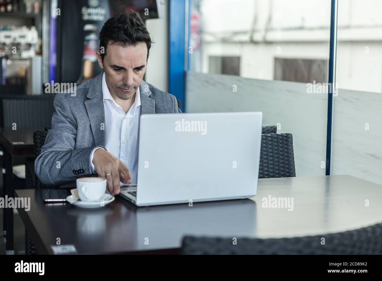 Homme d'affaires sérieux et concentré pour adultes, vêque d'une tenue habillée, assis à une table avec une tasse de café et un ordinateur portable tout en travaillant projet d'affaires dans moderne ca Banque D'Images