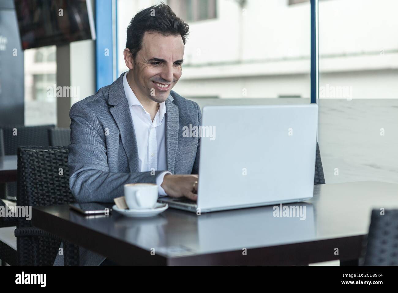 Manager homme adulte gai en tenue habillée assis à table avec une tasse de café et la lecture de bonnes nouvelles d'affaires sur ordinateur portable tout en ayant une pause dans moderne c Banque D'Images