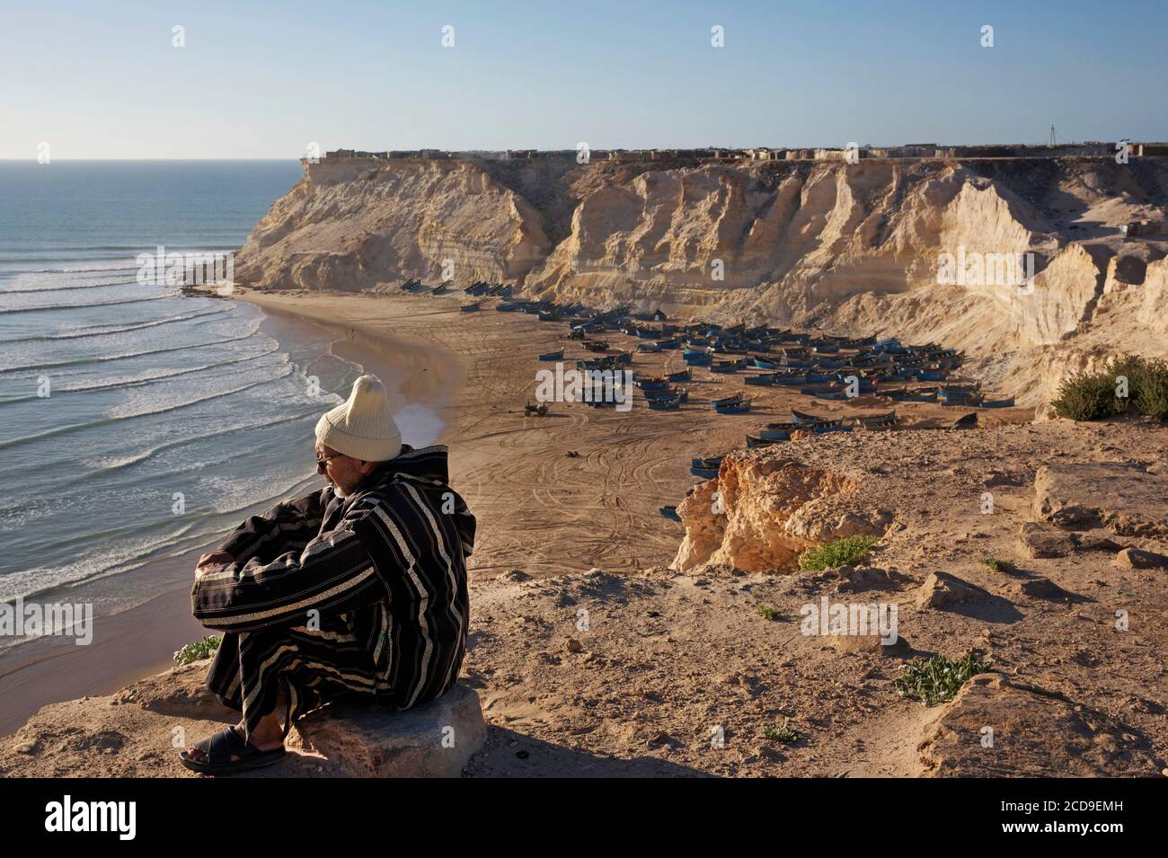 Maroc, Sahara occidental, Dakhla, vieil homme assis sur une falaise surplombant la plage d'Araiche et ses bateaux de pêche Banque D'Images