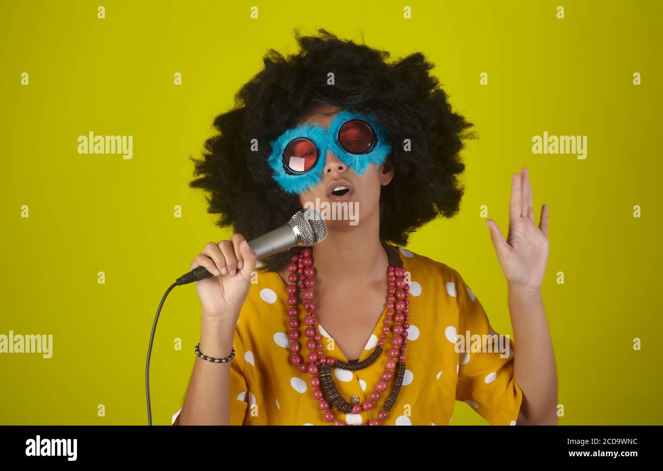 Jeune fille belle et souriante avec curly afro hairstyle et lunettes amusantes chantant avec microphone sur fond jaune Banque D'Images