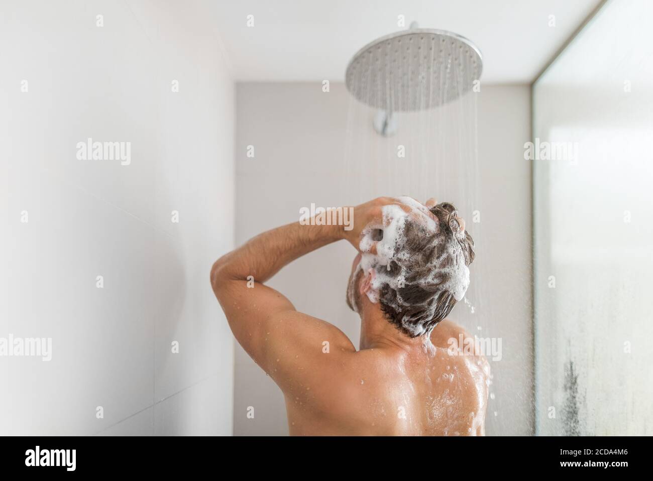 Homme prenant une douche lavant les cheveux sous l'eau tombant de la pomme de douche de pluie dans la salle de bain de luxe. Le style de vie de la jeune personne à la maison. Soins du corps Banque D'Images