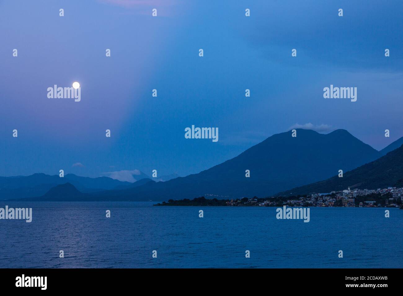 La pleine lune qui s'élève au-dessus du lac Atitlan au Guatemala. En premier plan se trouve la ville de San Pedro la Laguna. Les doubles sommets du dôme du volcan Toliman Banque D'Images