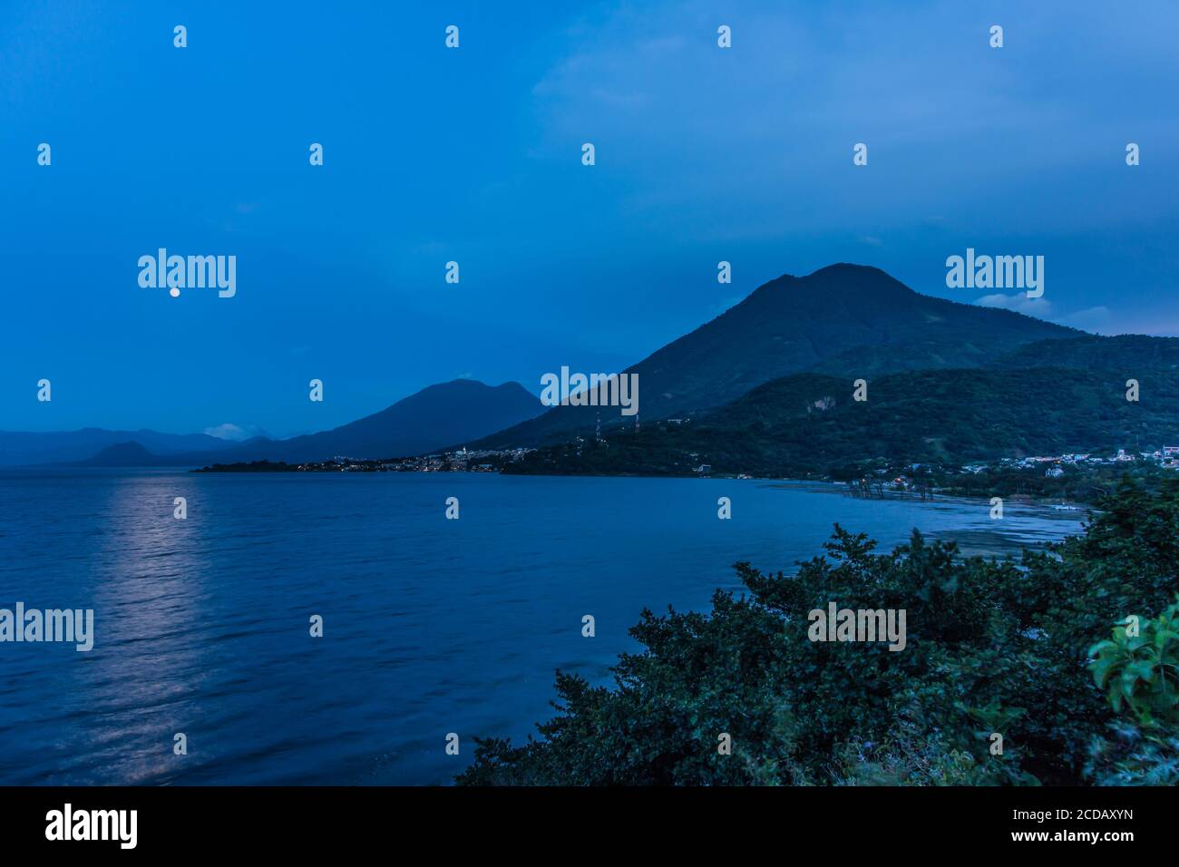 La pleine lune qui s'élève au-dessus du lac Atitlan au Guatemala. Au premier plan se trouve la ville de San Juan la Laguna. Les sommets doubles du volcan Toliman sont à Banque D'Images