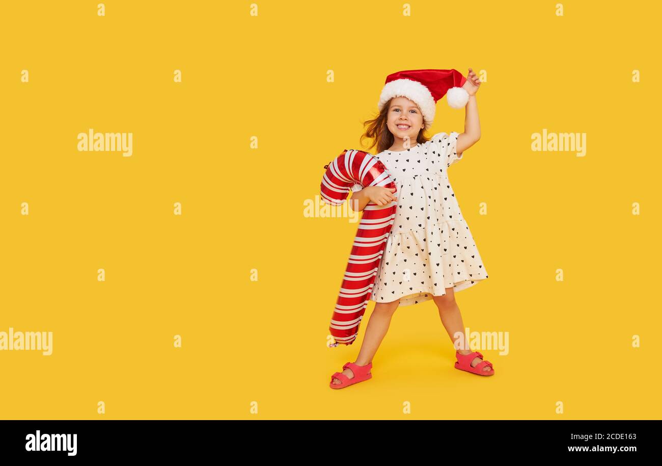 Une petite fille mignonne dans une robe de fantaisie et un chapeau de père Noël tient une forme gonflable d'une canne à sucre sur un fond jaune. 2021 vacances nouvel an con Banque D'Images