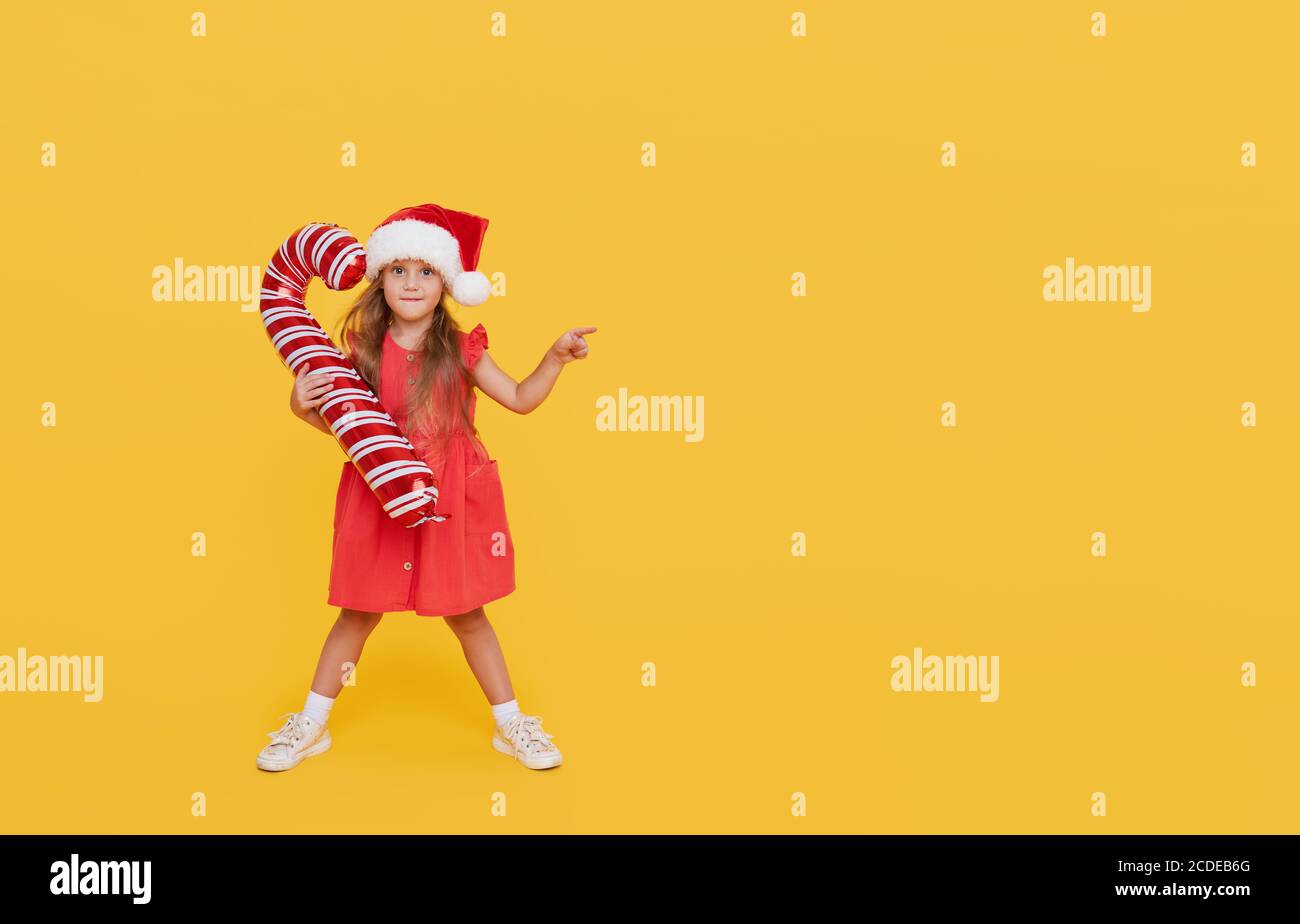 Une petite fille mignonne dans une robe de fantaisie et un chapeau de père Noël tient une forme gonflable d'une canne à sucre sur un fond jaune. 2021 vacances nouvel an con Banque D'Images