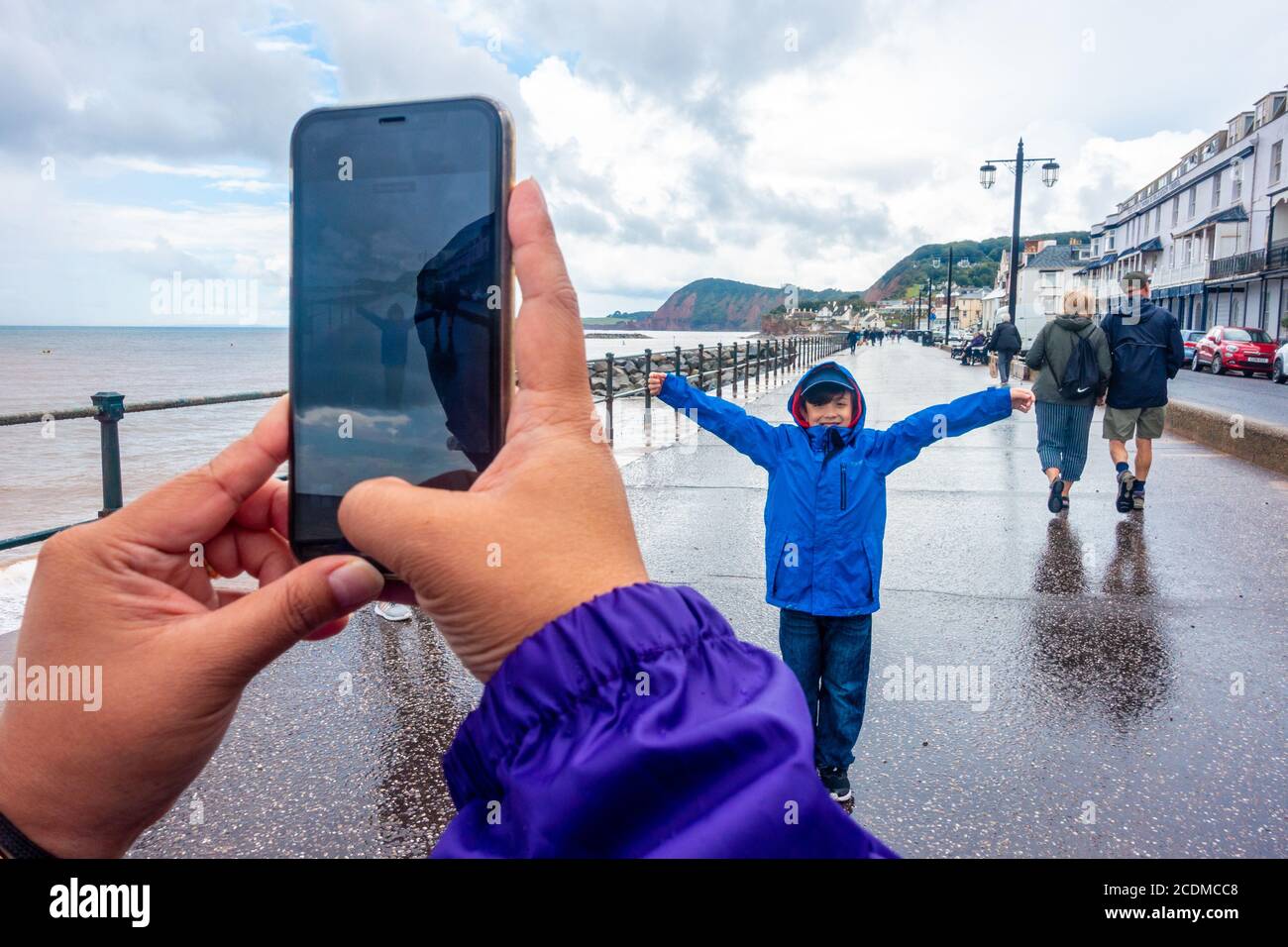 Gros plan d'une dame qui prend une photo de son fils à l'aide d'un téléphone portable sur l'esplanade, sur le front de mer, à Sidmouth, dans le sud du Devon, au Royaume-Uni, par une journée humide. Banque D'Images