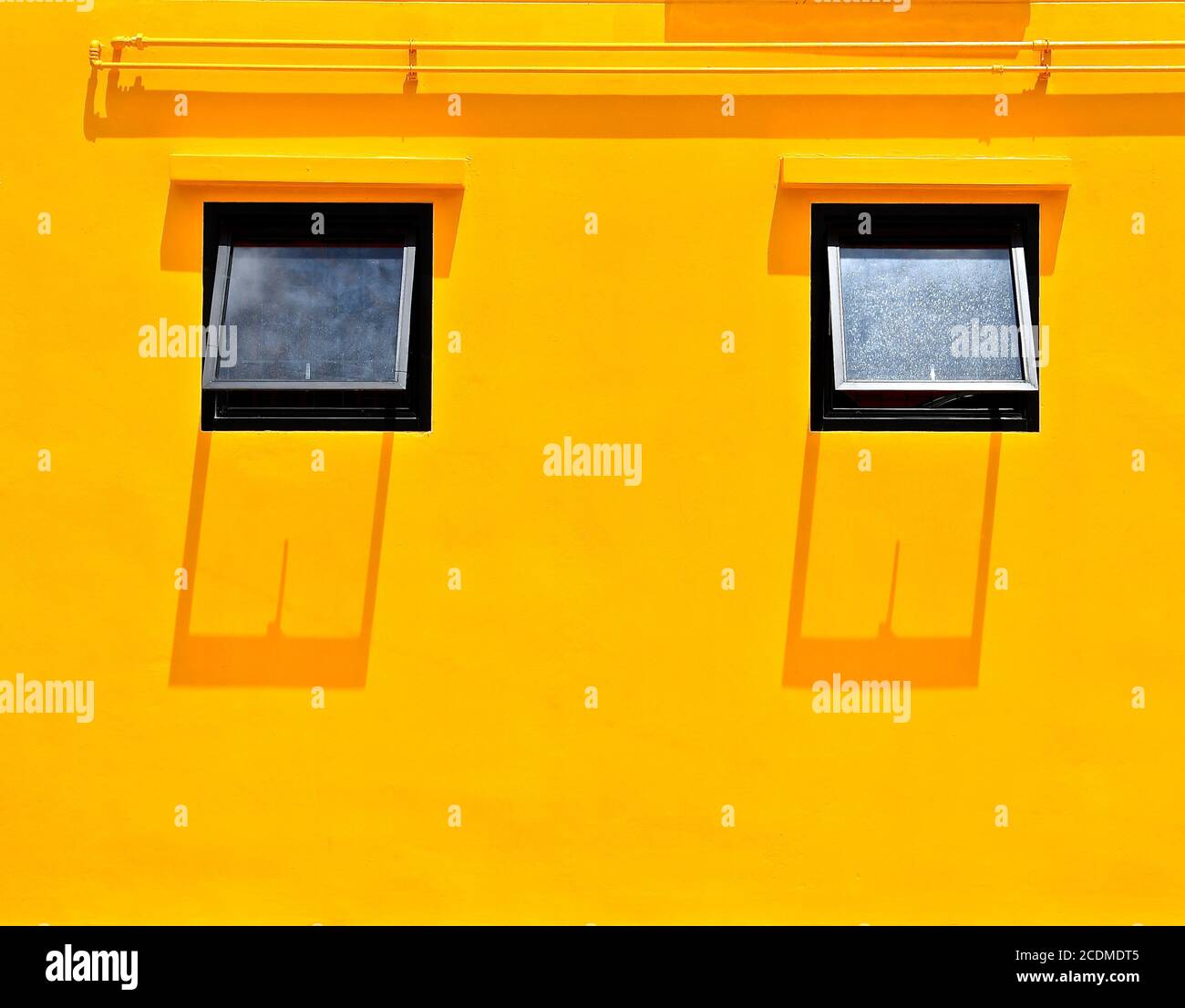 Un mur plâtré jaune vif Uni avec deux fenêtres ouvertes et des ombres projetées depuis le soleil de midi. Banque D'Images