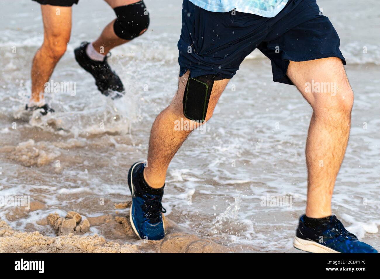 Les coureurs qui éclabousse dans l'océan en courant au bord de la plage avec un téléphone portable qui sort des shorts. Banque D'Images