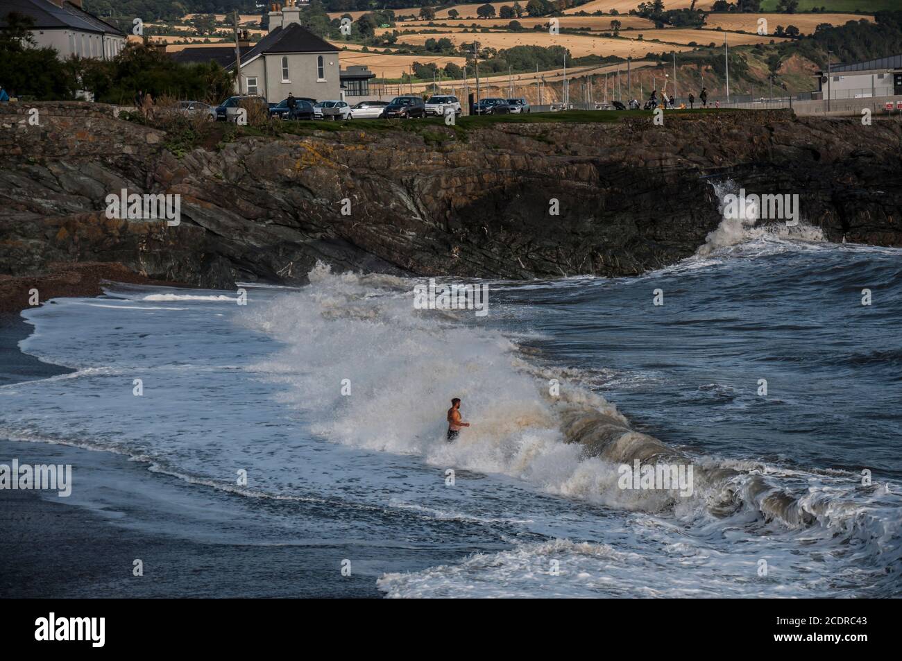 Visite de Greystones. L'homme de milieu d'âge reste devant la vague de rupture en profitant de ses éclaboussures. Banque D'Images