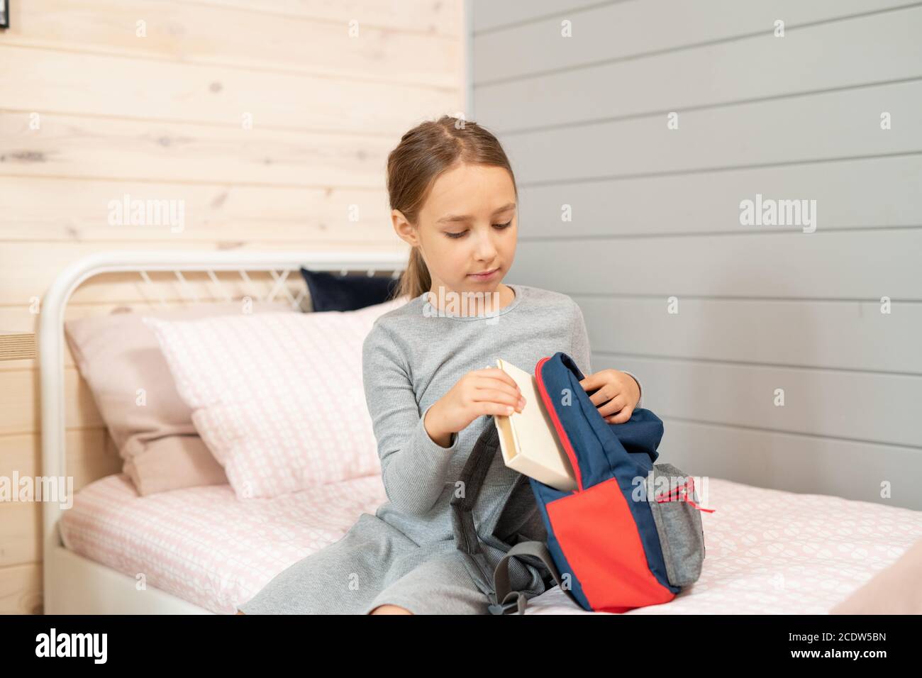 Grave mignonne écolière en robe grise mettant pile de livres dans un sac à dos Banque D'Images
