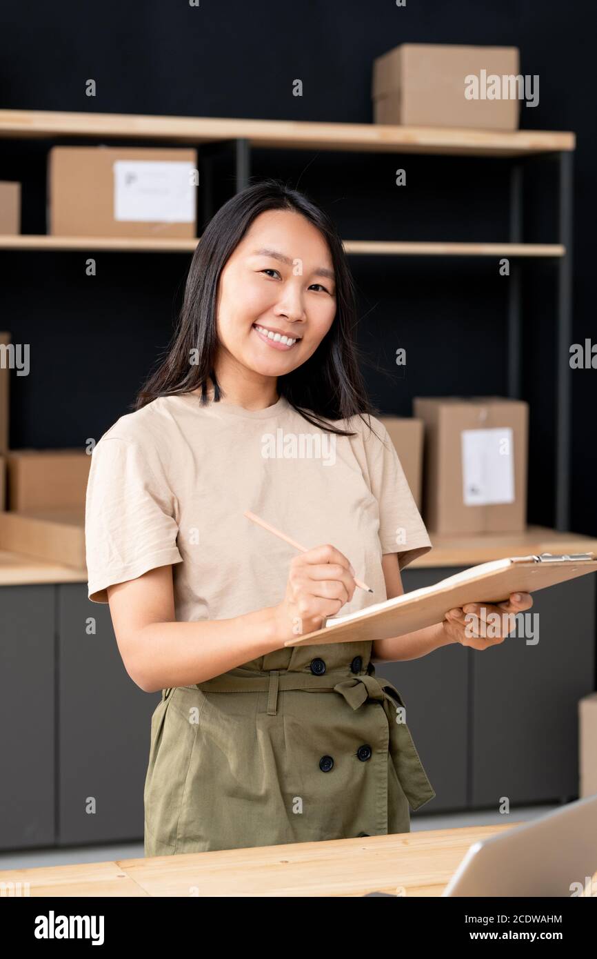 Bonne jeune femme asiatique avec des listes de vérification de stylo et de presse-papiers des commandes des clients Banque D'Images