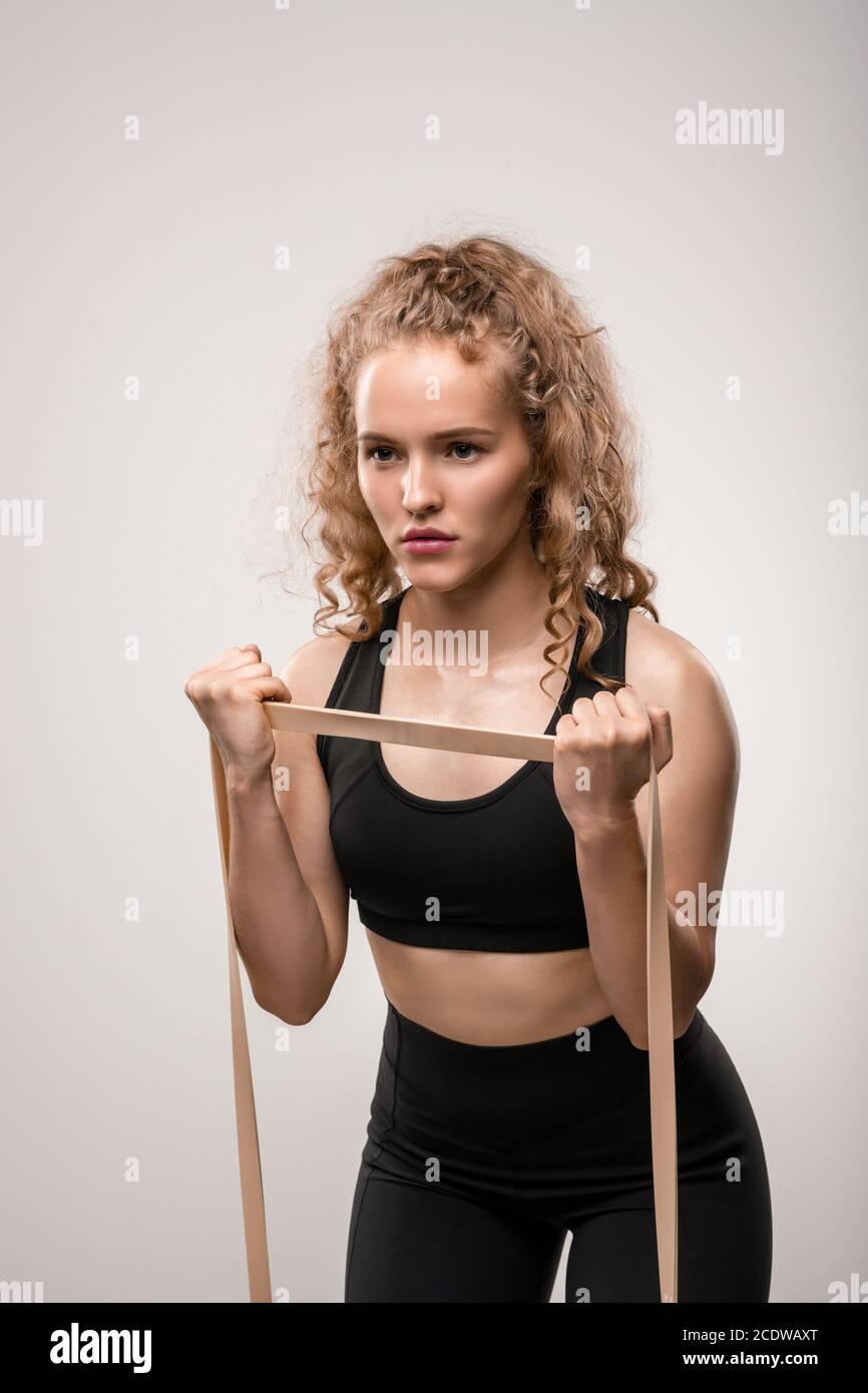 Jeune sportswoman avec cheveux blonds bouclés s'exerçant avec bande élastique dans la salle de sport Banque D'Images