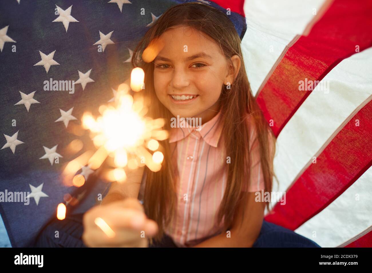 Portrait d'une jolie fille tenant des lumières étincelantes souriant à l'appareil photo contre l'arrière-plan du drapeau américain, espace de copie Banque D'Images