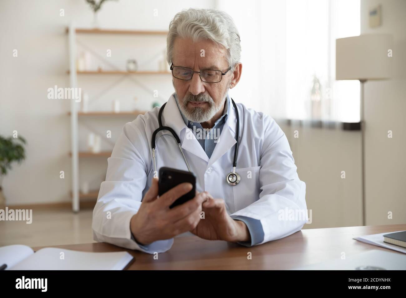 Médecin sérieux et mature regardant l'écran du smartphone, assis au bureau Banque D'Images