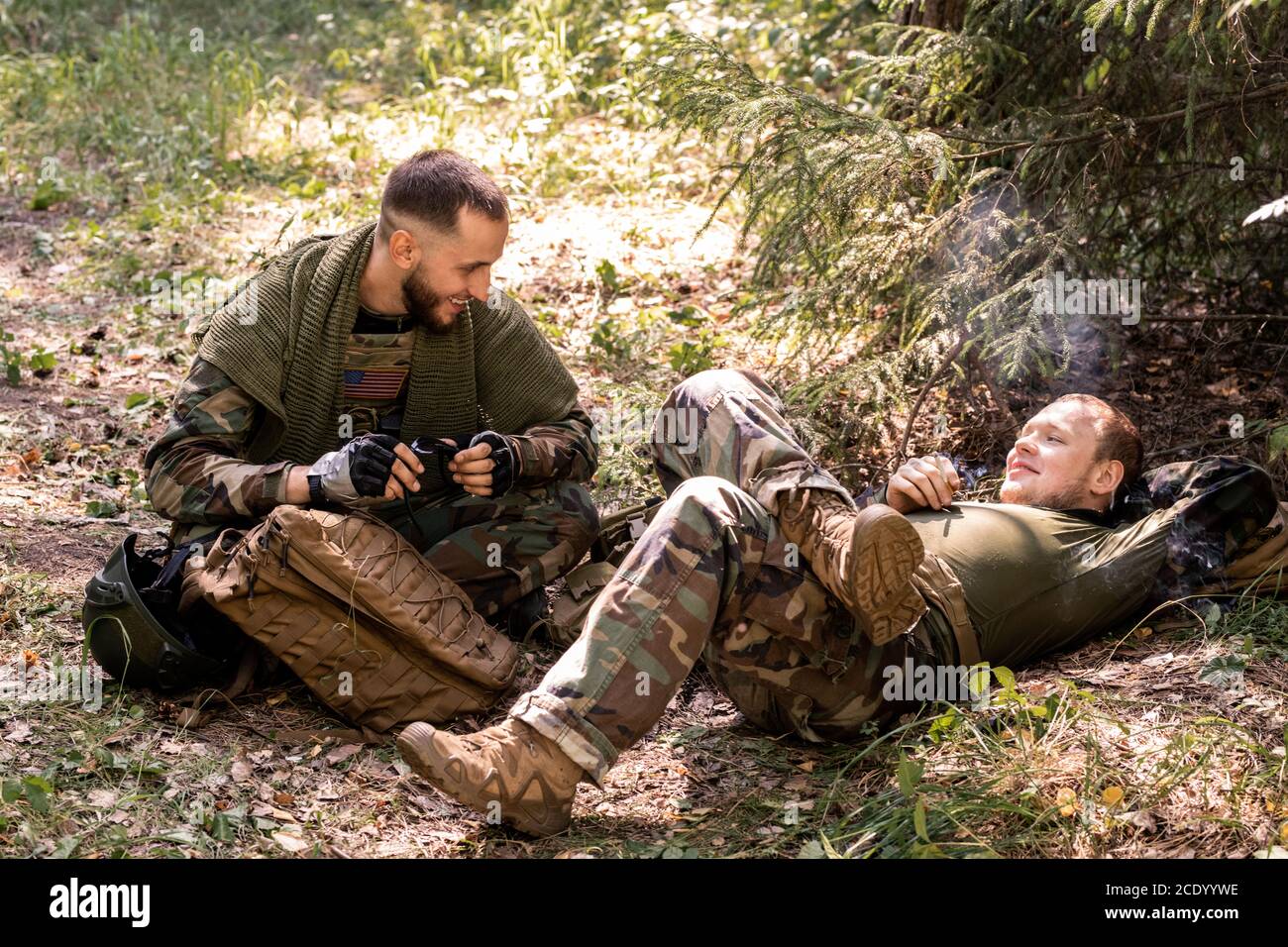 De jeunes soldats positifs en uniforme militaire fument de la cigarette et bavardent tout en se reposant dans un abri forestier Banque D'Images