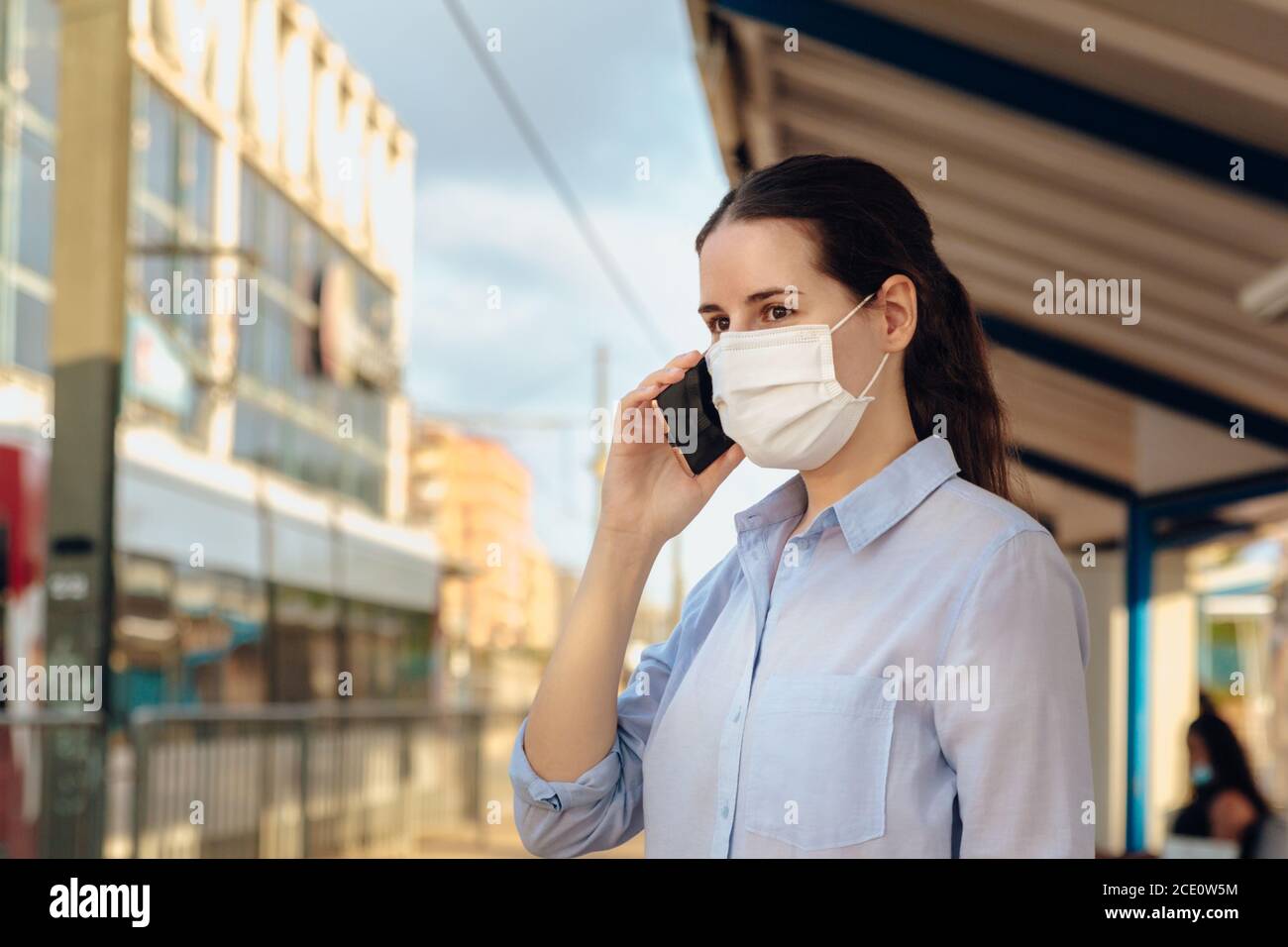 Photo d'une jeune femme portant un masque et parlant au téléphone en attendant les transports en commun. Nouvelle normale Banque D'Images