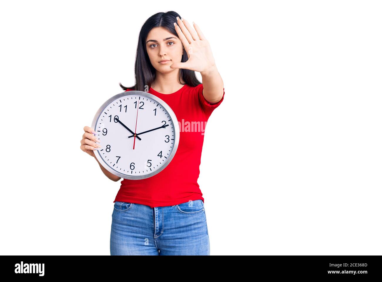 Jeune belle fille tenant horloge avec main ouverte faisant signe d'arrêt avec expression sérieuse et confiante, geste de défense Banque D'Images