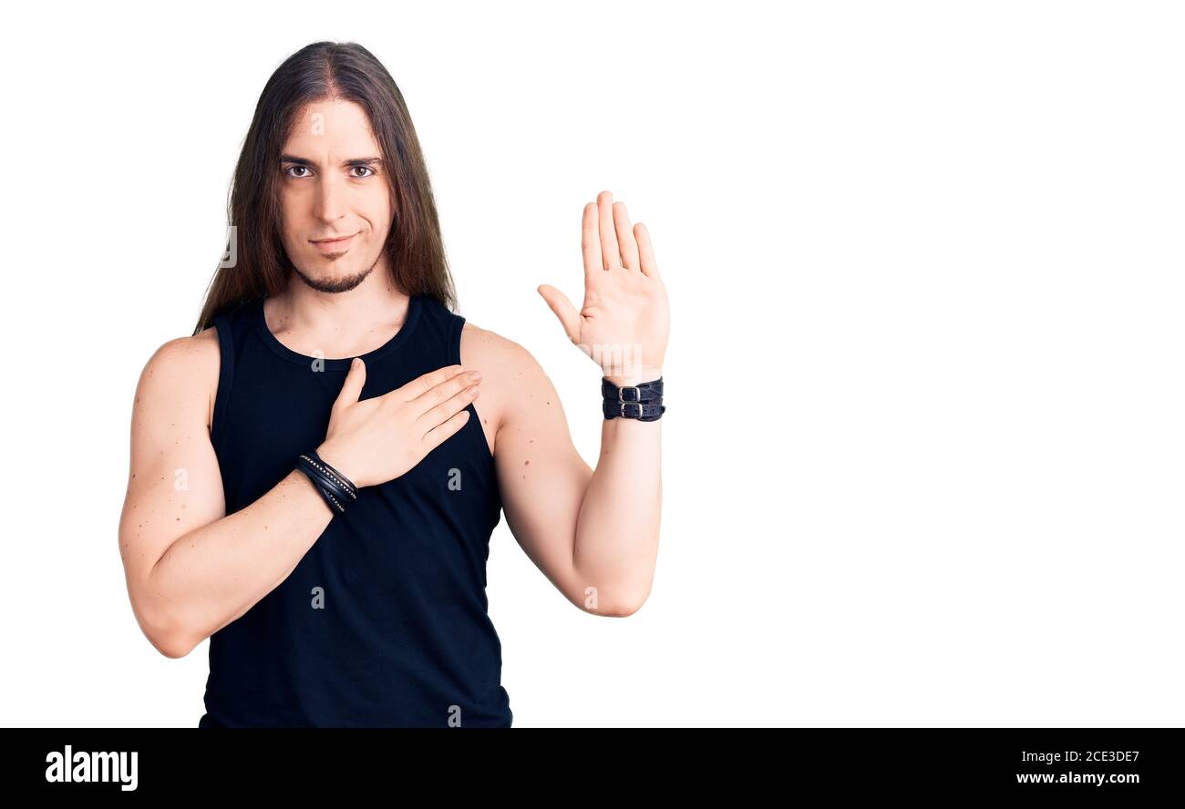 Jeune homme adulte aux cheveux longs portant un style de goth avec des vêtements noirs serrant avec la main sur la poitrine et la paume ouverte, faisant un serment de loyauté promesse Banque D'Images