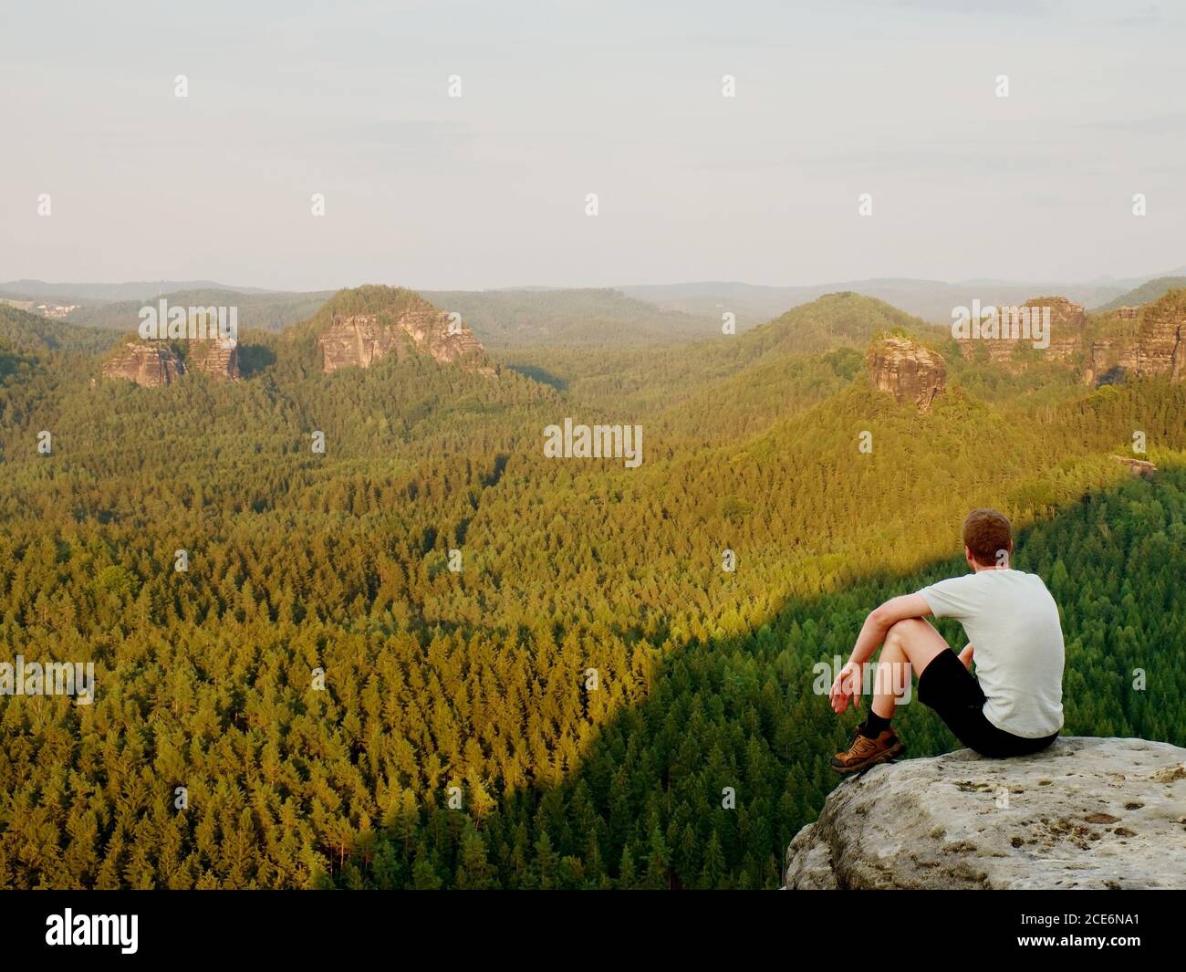 Moment de solitude. Un homme en chemise s'assoit sur la falaise de roche et regarde dans la vallée de la forêt. Banque D'Images
