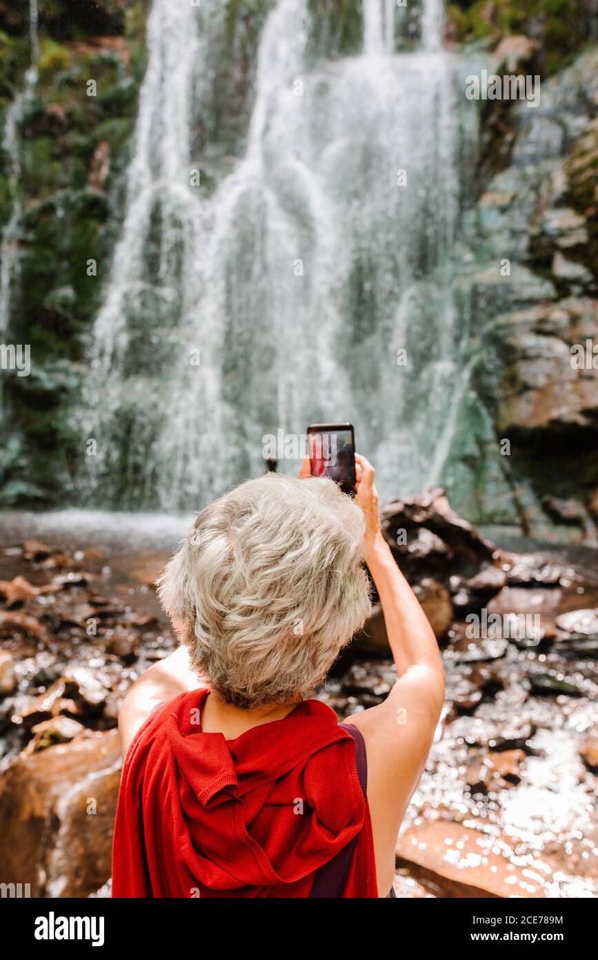 Vue arrière de la touriste féminine sans visage avec des cheveux gris utilisant smartphone et prendre des photos de la magnifique chute d'eau pendant l'aventure estivale Banque D'Images