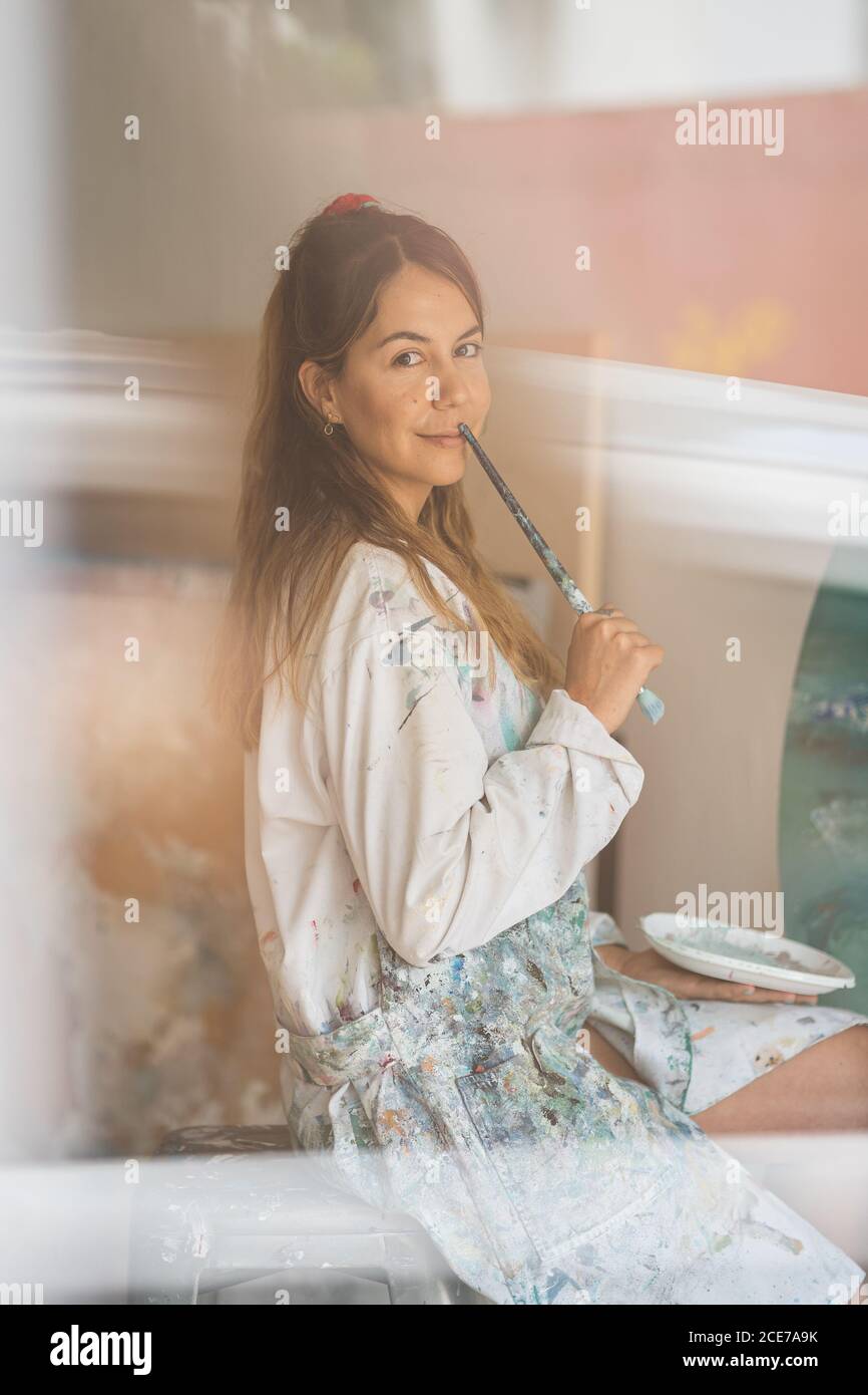 Vue latérale d'une jeune femme talentueuse dans des vêtements salissants avec un pinceau et une palette souriant et regardant l'appareil photo pendant assis devant le chevalet et dessin d'une photo sur toile en studio créatif Banque D'Images
