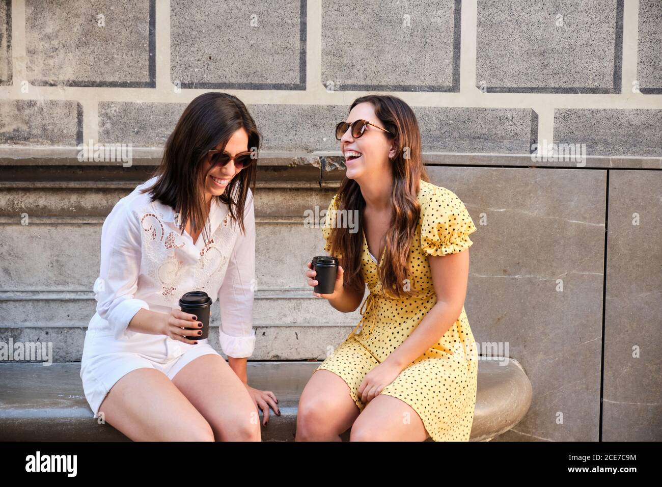 Femmes amicales en été, assis sur un banc dans la rue avec un café à emporter et rire ensemble à la plaisanterie Banque D'Images
