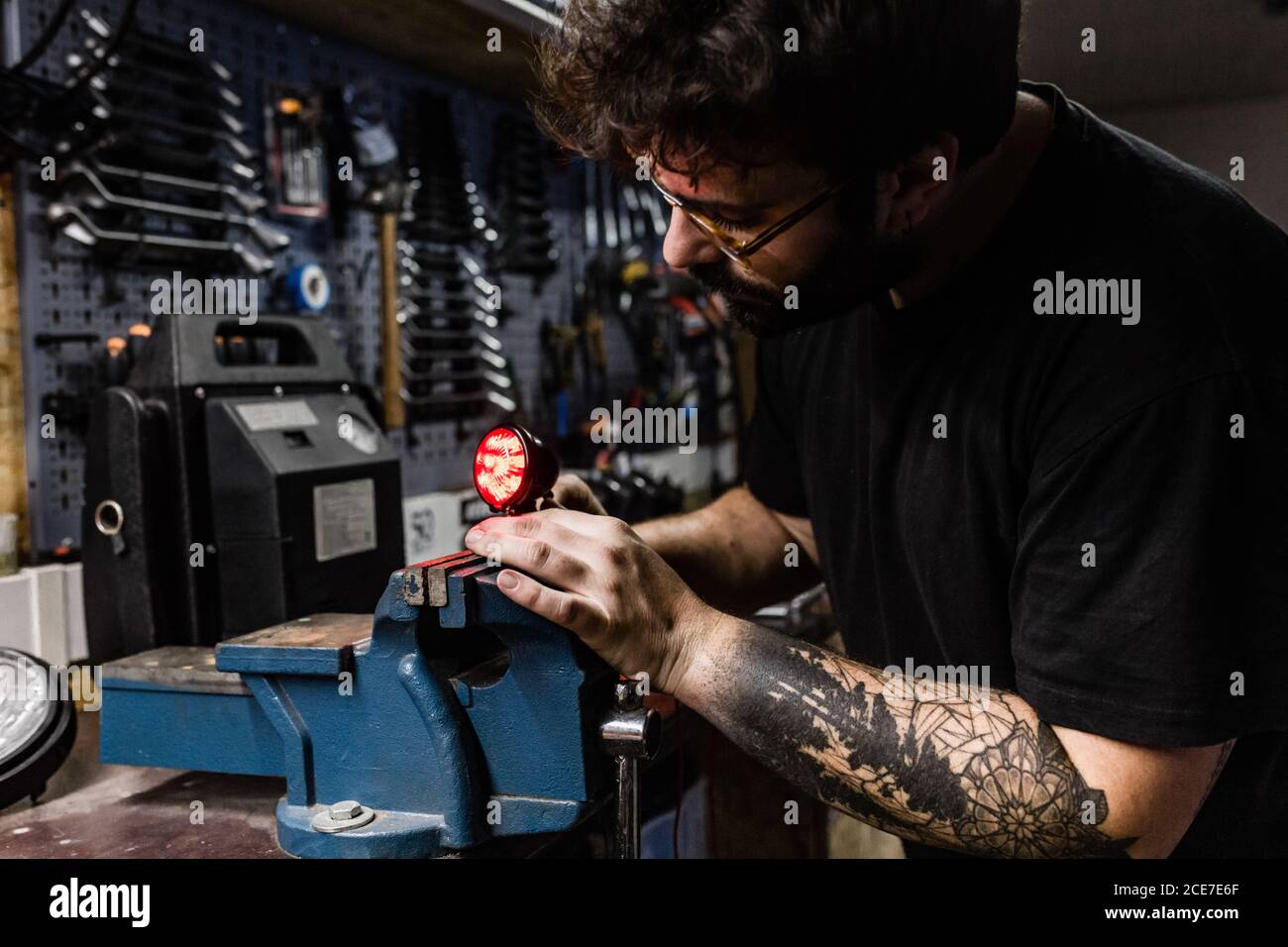 Vue latérale d'un mécanicien masculin concentré avec tatouages à l'aide d'un étau et réparer le feu arrière rouge de la moto pendant le travail atelier Banque D'Images Vue latérale d'un mécanicien masculin concentré avec tatouages à l'aide d'un étau et réparer le feu arrière rouge de la moto pendant le travail atelier Banque D'Images