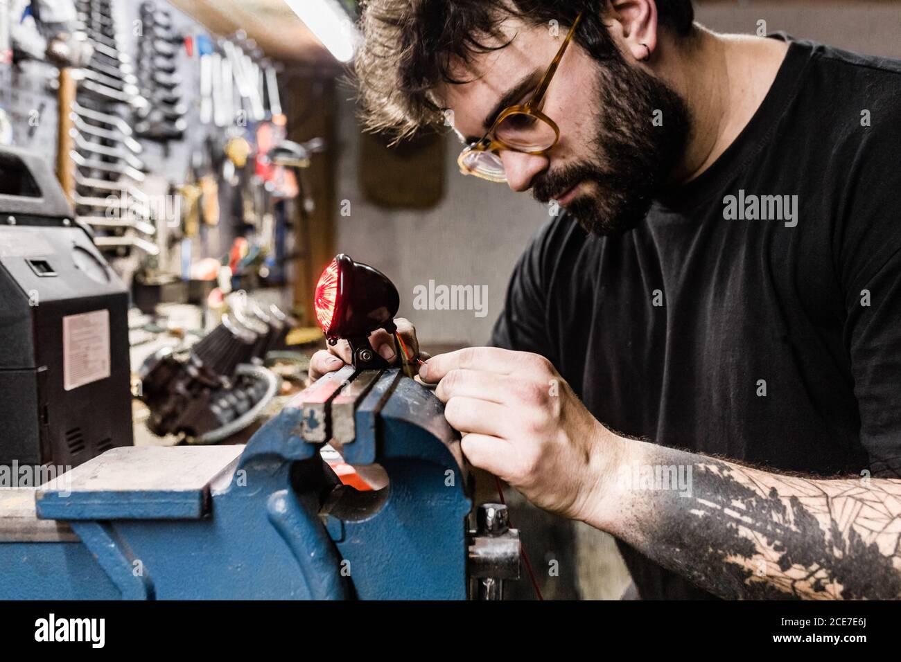Vue latérale d'un mécanicien masculin concentré avec tatouages à l'aide d'un étau et réparer le feu arrière rouge de la moto pendant le travail atelier Banque D'Images Vue latérale d'un mécanicien masculin concentré avec tatouages à l'aide d'un étau et réparer le feu arrière rouge de la moto pendant le travail atelier Banque D'Images