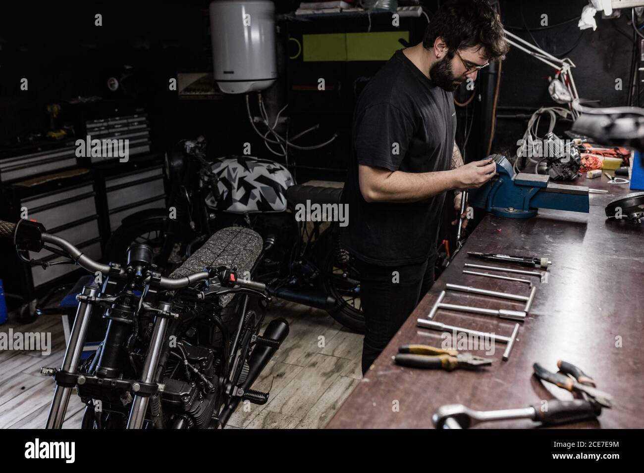 Vue latérale d'un mécanicien masculin concentré avec tatouages à l'aide d'un étau et réparer le feu arrière rouge de la moto pendant le travail atelier Banque D'Images Vue latérale d'un mécanicien masculin concentré avec tatouages à l'aide d'un étau et réparer le feu arrière rouge de la moto pendant le travail atelier Banque D'Images
