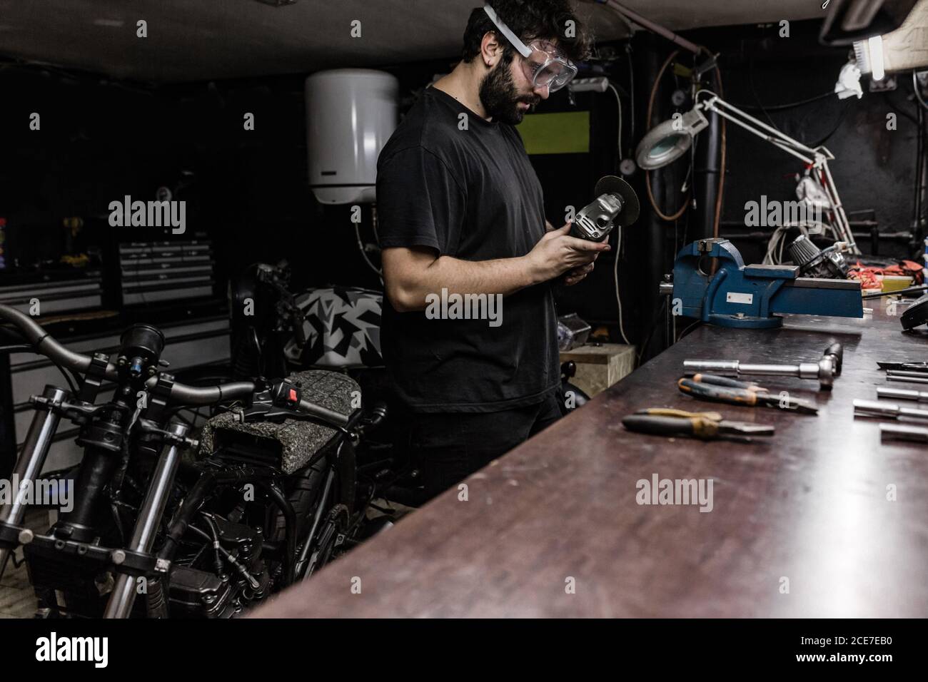 Vue latérale d'un homme maître barbu portant des lunettes de protection debout dans un atelier de travail de moindre qualité et découpe des détails métalliques avec une meuleuse Banque D'Images