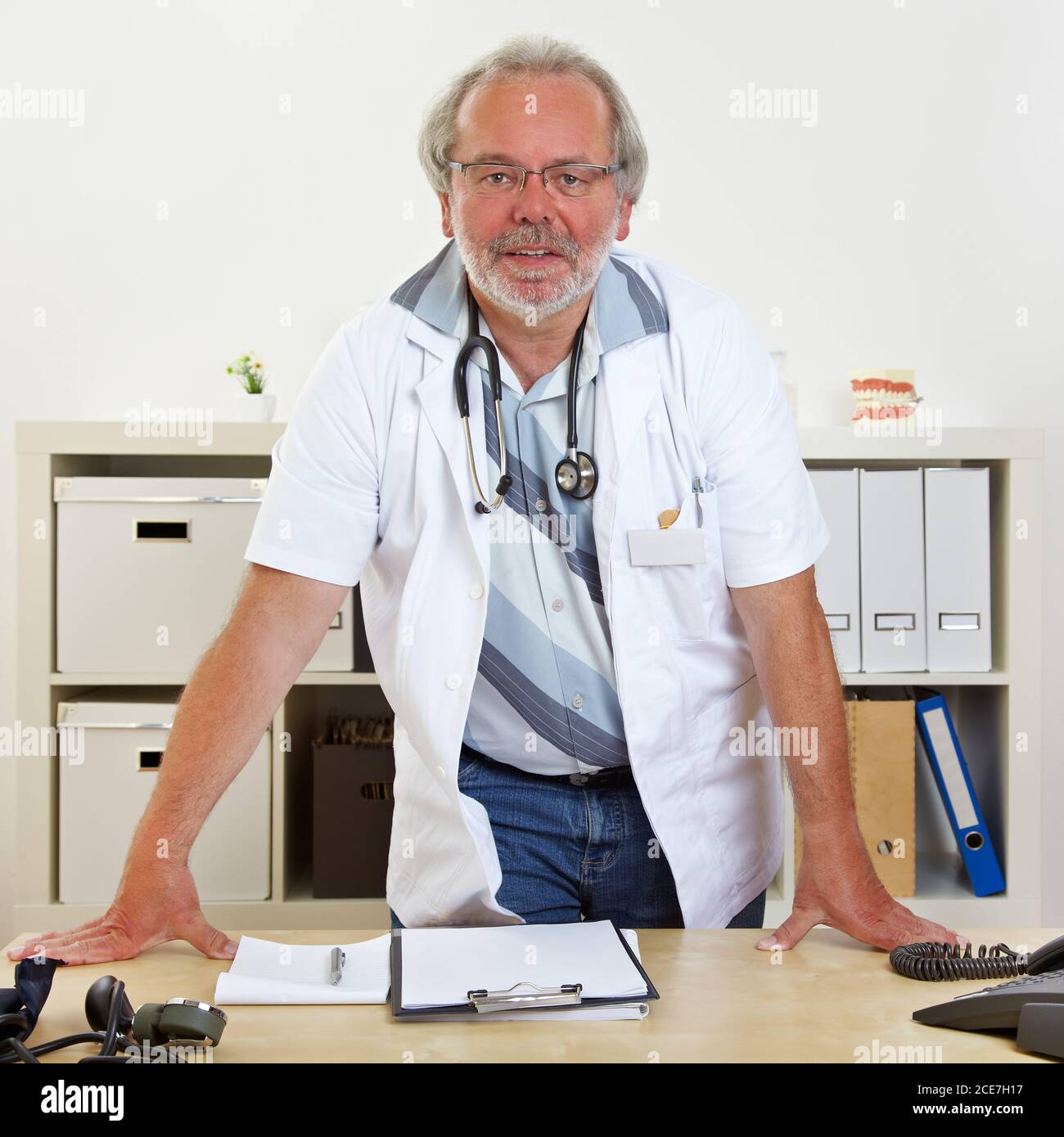 Le médecin âgé est debout derrière son bureau dans celui du médecin bureau Banque D'Images