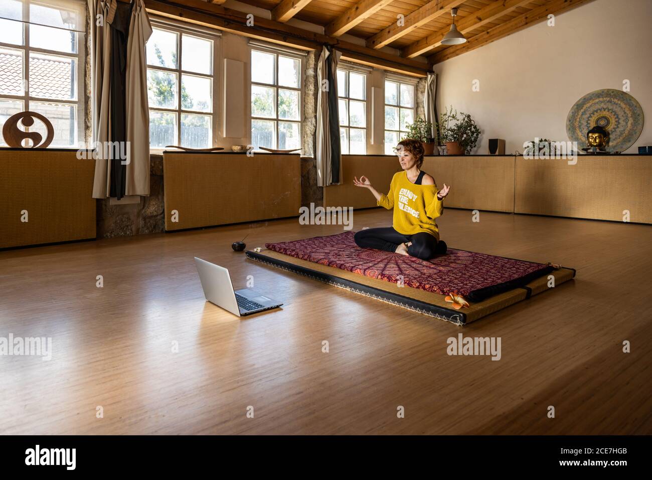 Femme calme dans les vêtements de sport assis sur un tapis dans un studio spacieux Et pratiquer le yoga à Padmasana tout en filmant un tutoriel en ligne sur ordinateur portable Banque D'Images