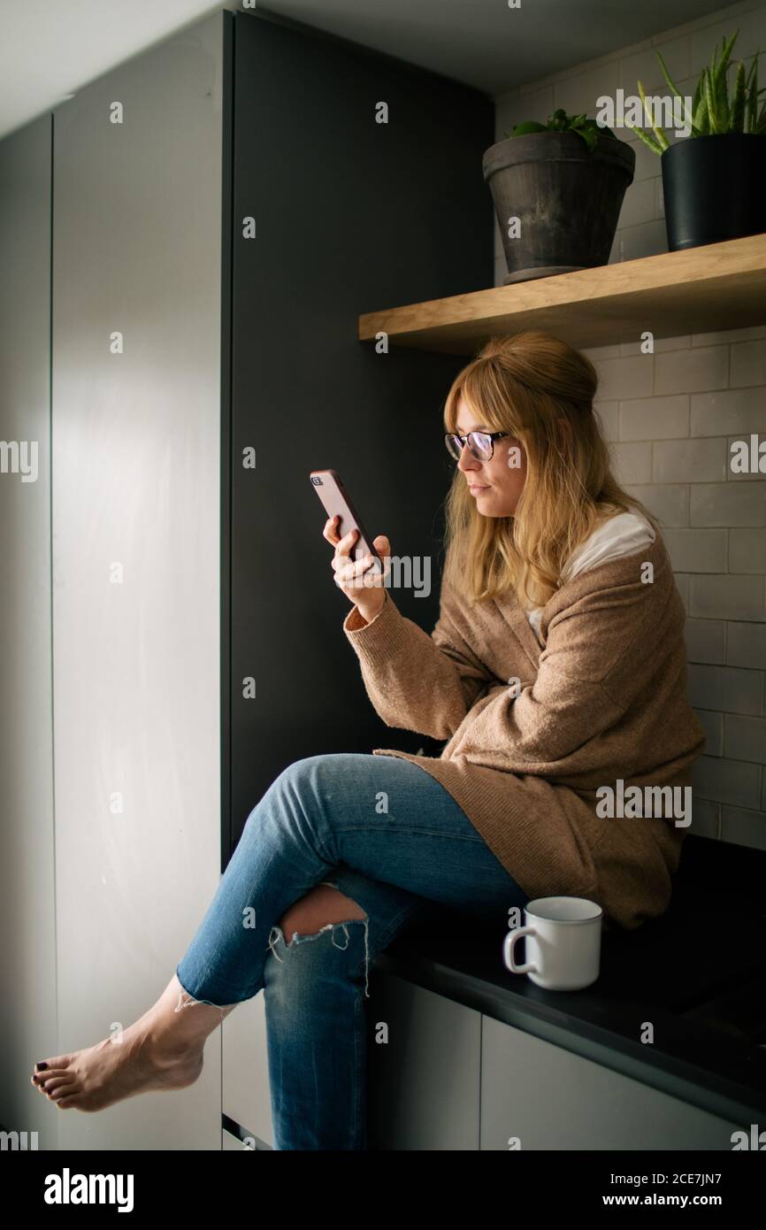Femme sereine dans des vêtements confortables assis sur le comptoir dans la cuisine et lire des messages sur les réseaux sociaux tout en dégustant une boisson fraîche le matin Banque D'Images