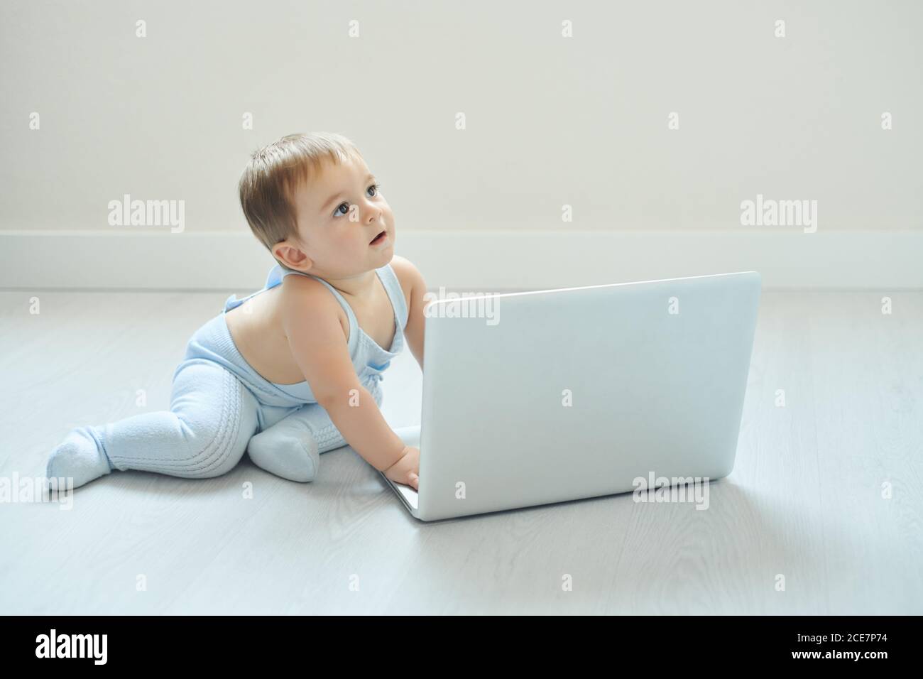 Vue latérale de Happy Little tout-petit assis sur le sol et appuyez sur les touches de l'ordinateur portable tout en jouant seul dans une pièce lumineuse à la maison Banque D'Images