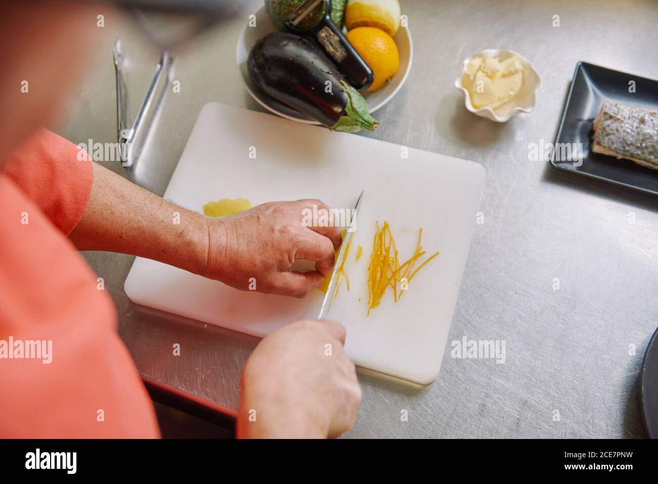 Du dessus de la récolte anonyme chef coupant le zeste de citron frais pendant la cuisson, près de la plaque avec du poisson et des légumes crus sur une surface métallique Banque D'Images