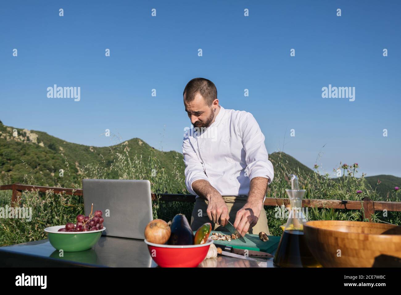 Chef masculin concentré dans la recette de lecture uniforme sur ordinateur portable et couper des légumes tout en cuisinant le déjeuner sur la terrasse extérieure du restaurant en campagne Banque D'Images