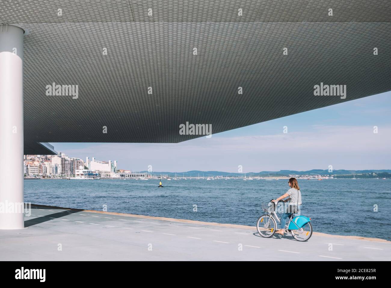 Vue latérale de la femme en tenue décontractée à vélo près bâtiment moderne et front de mer pendant la journée ensoleillée Banque D'Images