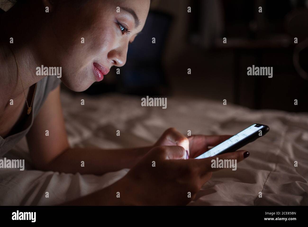 Vue latérale d'une jeune femme asiatique joyeuse utilisant un smartphone pendant couché dans le lit la nuit à la maison Banque D'Images