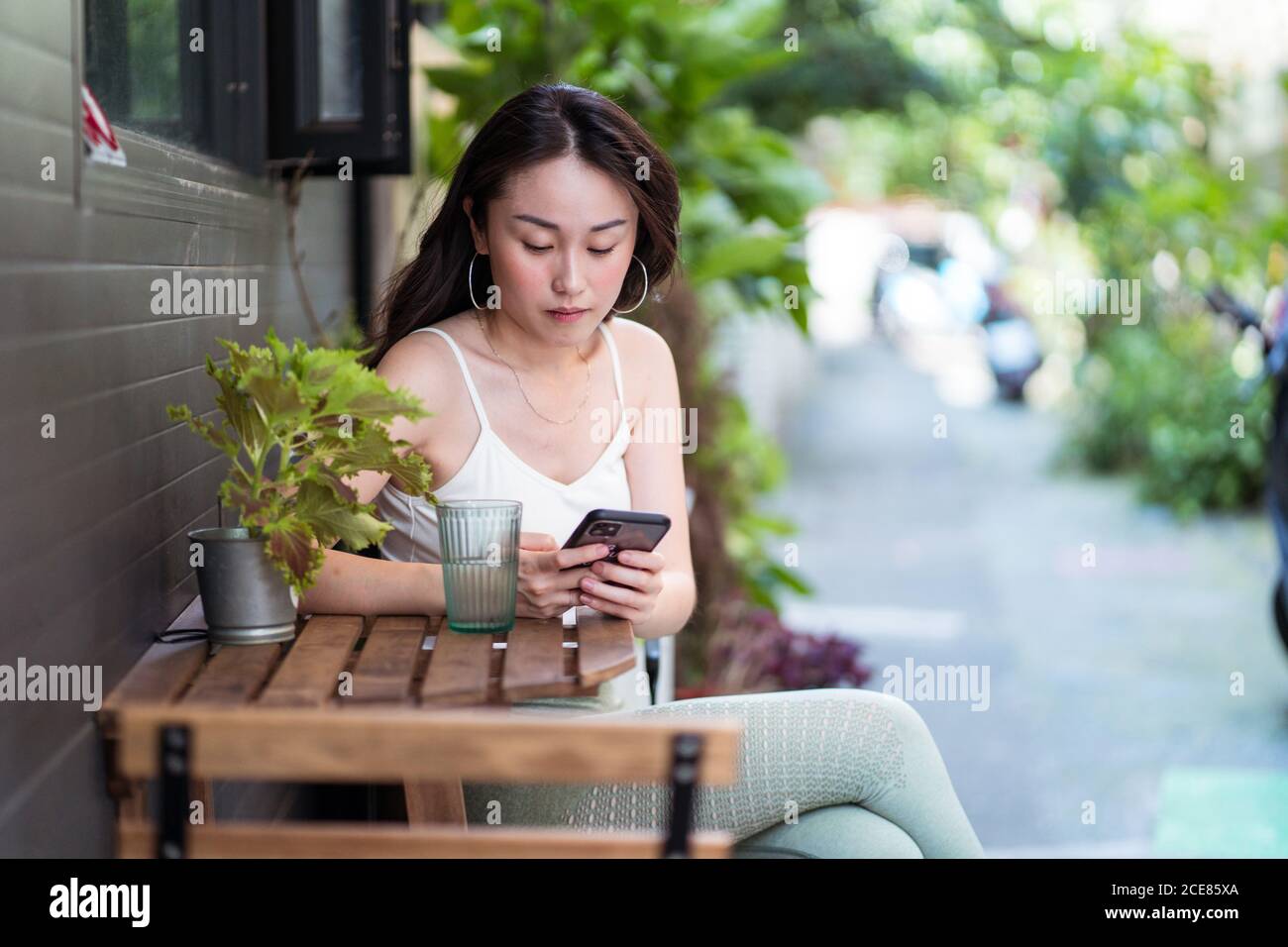 Jeune femme ethnique en tenue décontractée assise avec des jambes croisées à une table en bois avec un verre d'eau près de la maison cour tout en surfant sur internet sur un téléphone portable dans la lumière du jour Banque D'Images