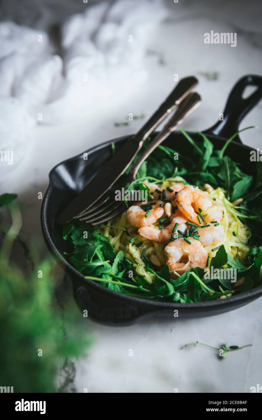 De la portion ci-dessus de délicieuses nouilles courgettes aux crevettes bouillies et des feuilles d'épinards fraîches placées sur la poêle avec la fourchette et couteau sur table en marbre Banque D'Images