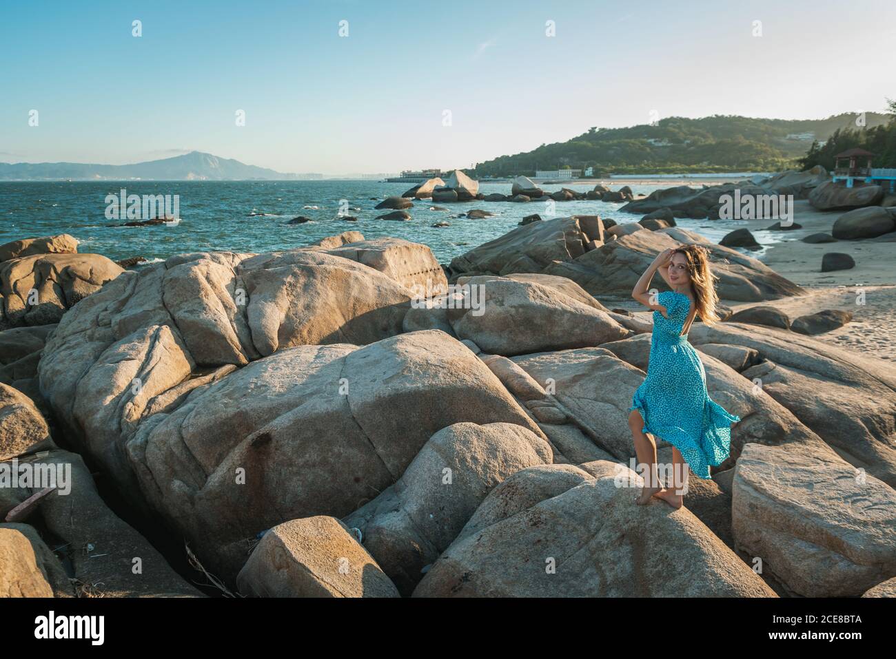 Corps complet de jeune femme barefooted touriste en bleu clair habillez-vous sur une immense pierre sur une plage de sable et profitez vue pittoresque sur la mer au coucher du soleil Banque D'Images