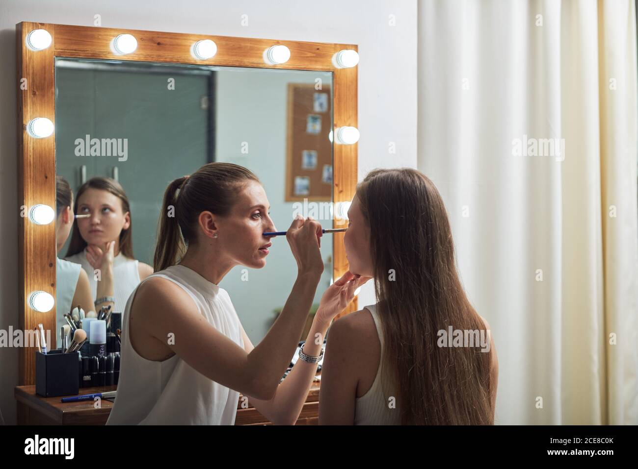 Jeune femme positive maquilleuse appliquant de la poudre sur le visage des clients en utilisant la brosse contre grand miroir confortable avec lumières dedans salon de beauté contemporain Banque D'Images