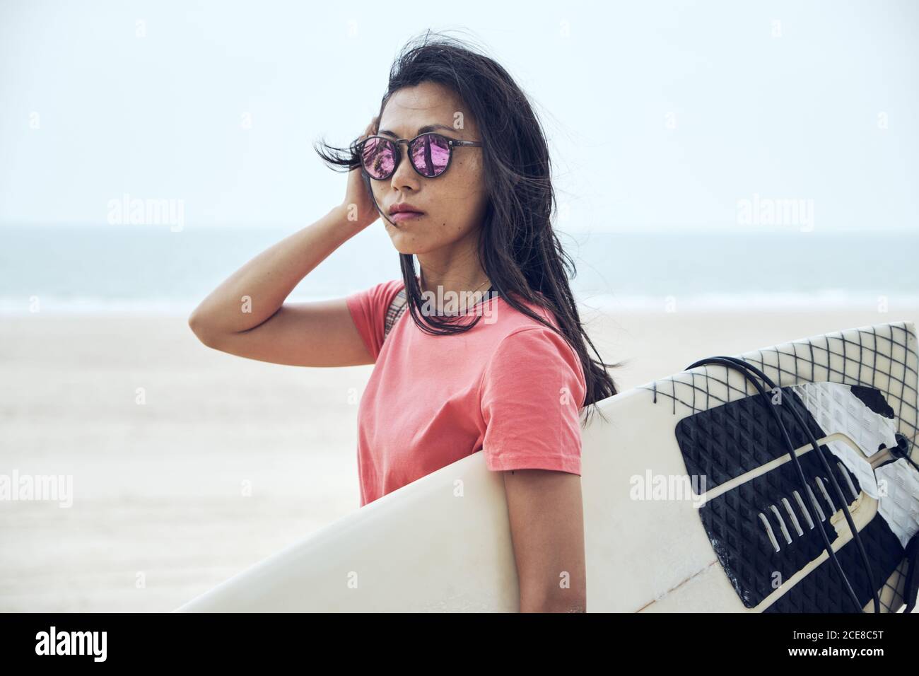 Jeune femme asiatique surfeuse en tenue d'été marchant sur du sable plage et transport de planches de surf contre mer bleu calme regardant appareil photo Banque D'Images