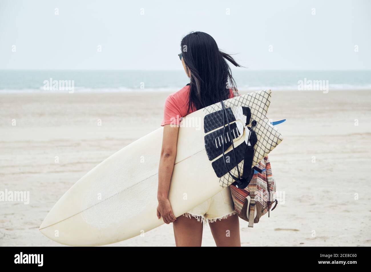 Vue arrière d'une jeune femme de surf méconnaissable en tenue estivale marcher sur une plage de sable et porter une planche de surf contre un bleu calme vue sur la mer Banque D'Images